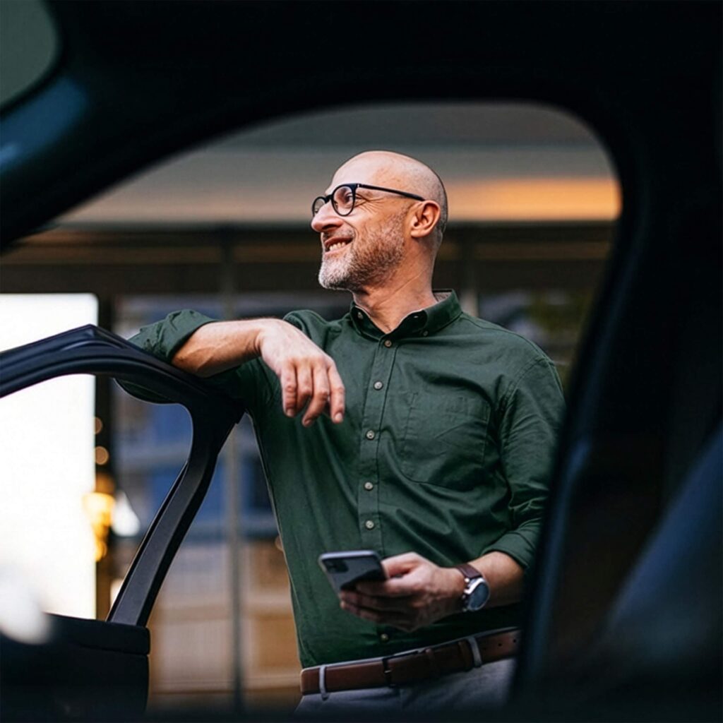 Un homme se tient avec son mobile à la main près d'une voiture et regarde au loin.