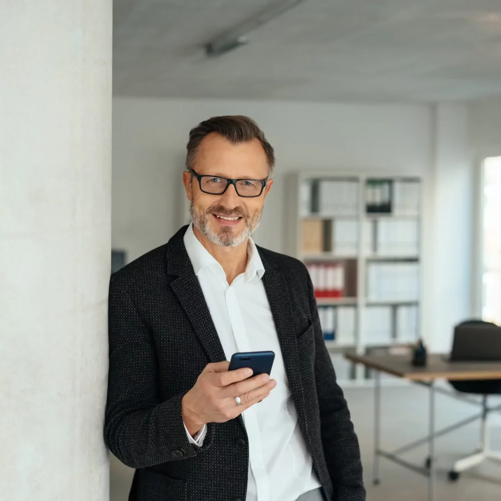 Homme d’affaires élégant tenant son téléphone mobile alors qu’il se détend appuyé contre un mur intérieur de bureau en souriant à la caméra